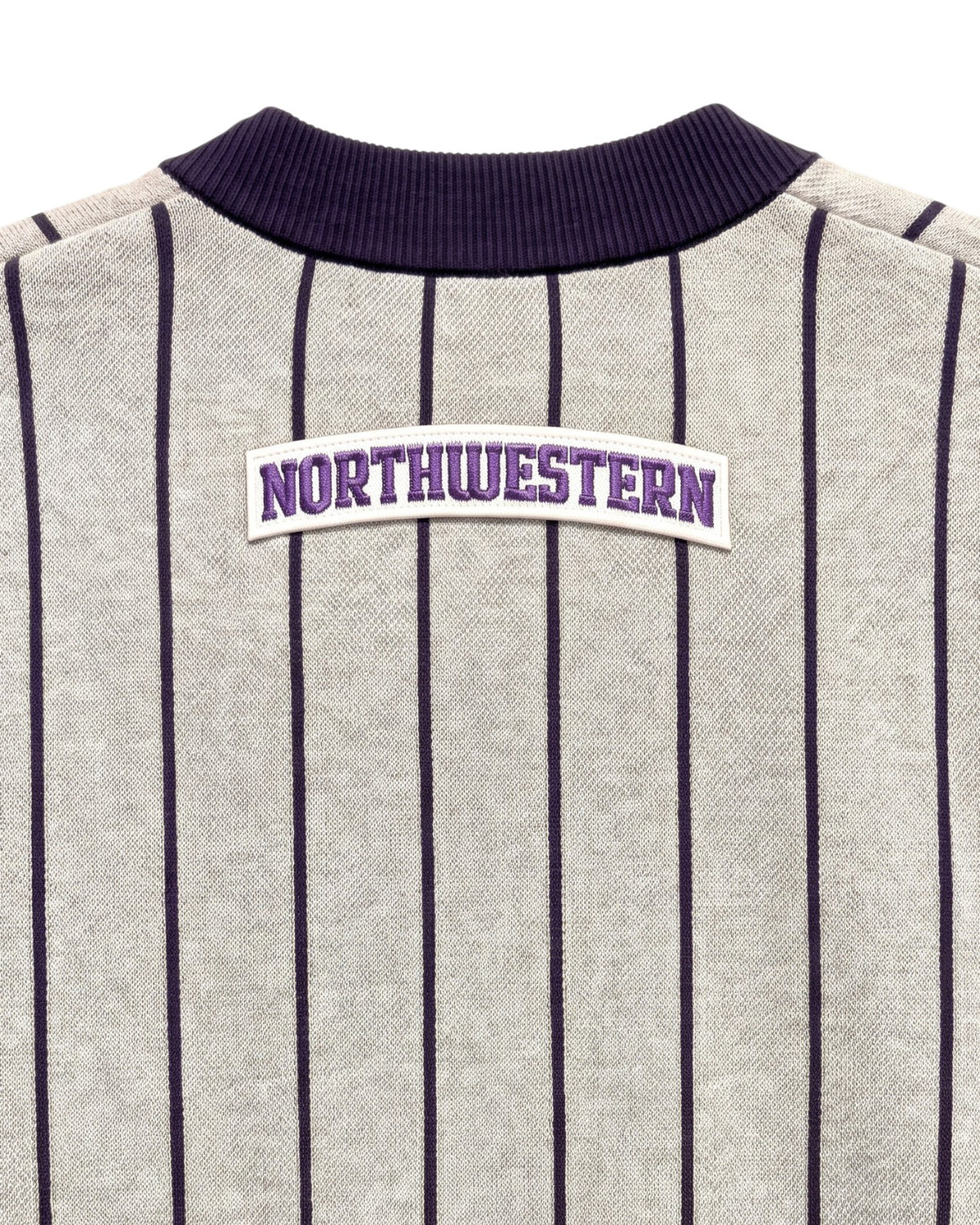 Close up of light grey and purple Northwestern University heritage pinstripe classic crewneck knit sweater with arched white varsity patch with NORTHWESTERN embroidered in purple sewn onto the upper center back below the collar.