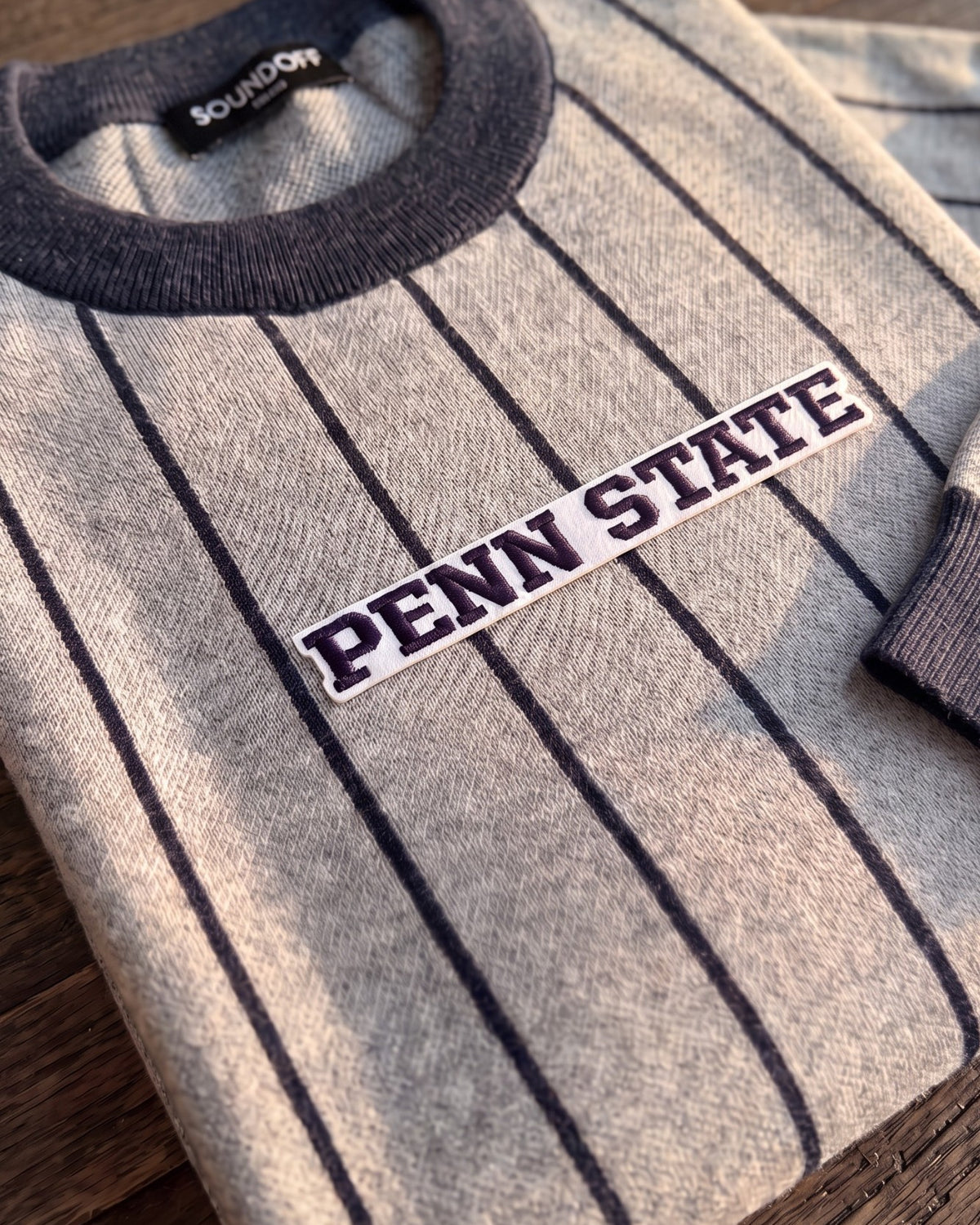 Penn State University block letter embroidered iron-on varsity patch in navy and white sewn onto center front chest of light grey and navy pinstripe classic crewneck knit sweater