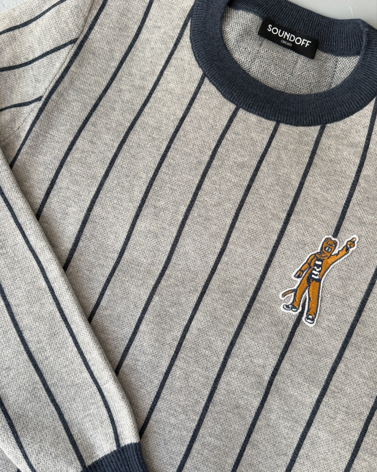 Penn State Nittany Lion mascot embroidered crest patch in brown, navy and white sewn onto left chest of light grey classic crewneck knit sweater with navy pinstripes and trim.