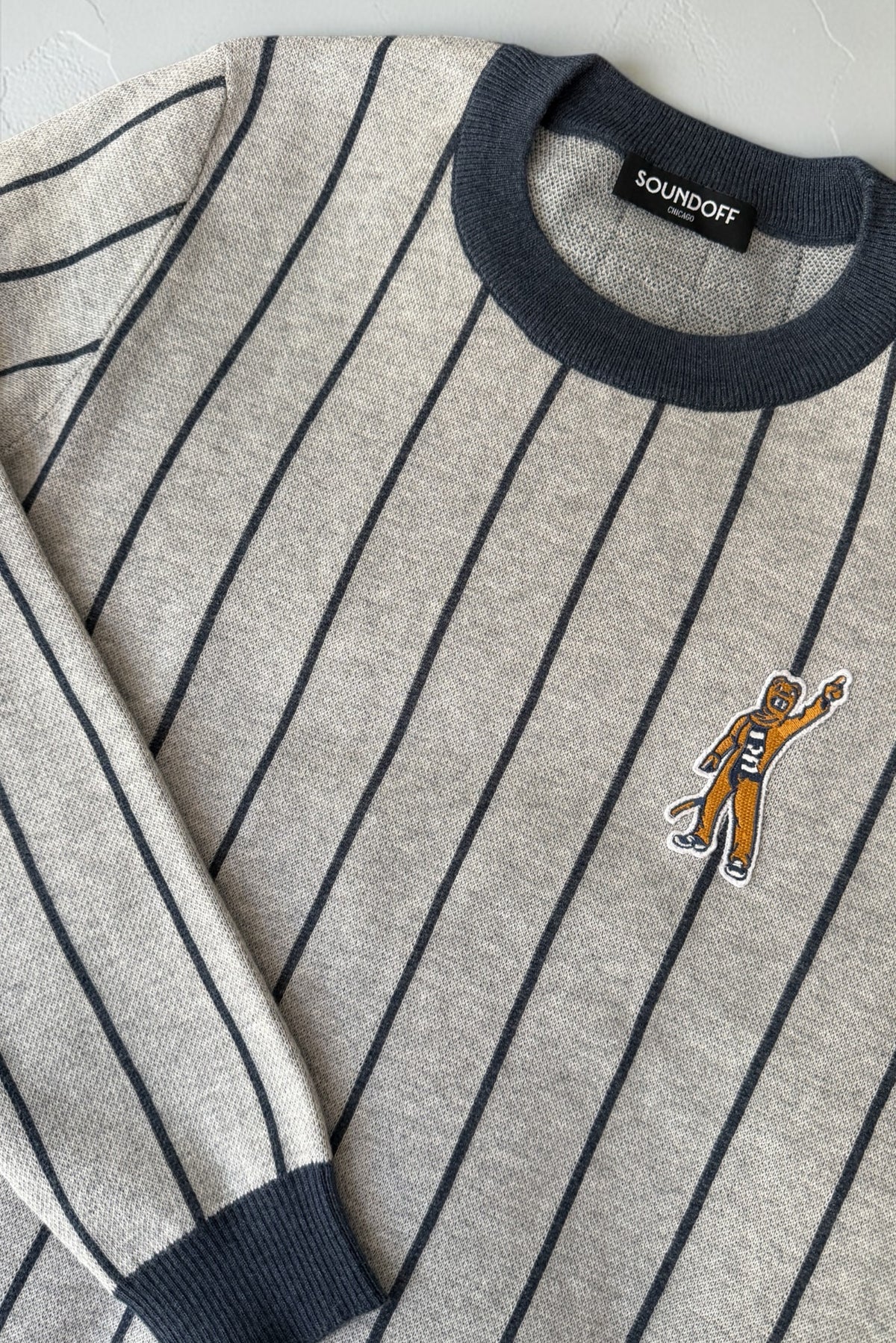 Penn State Nittany Lion mascot embroidered crest patch in brown, navy and white sewn onto left chest of light grey classic crewneck knit sweater with navy pinstripes and trim.