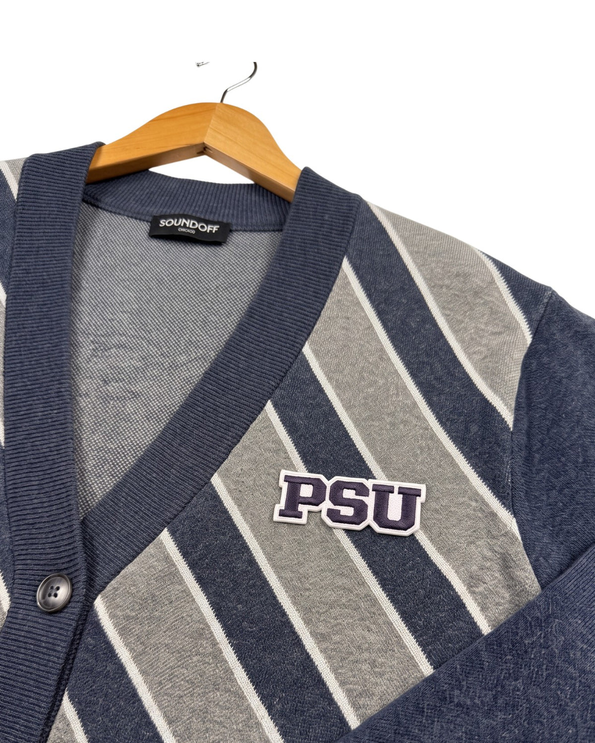 Penn State PSU crest embroidered patch in navy and white sewn onto left chest of navy cardigan knit sweater with white and light grey repp stripes.