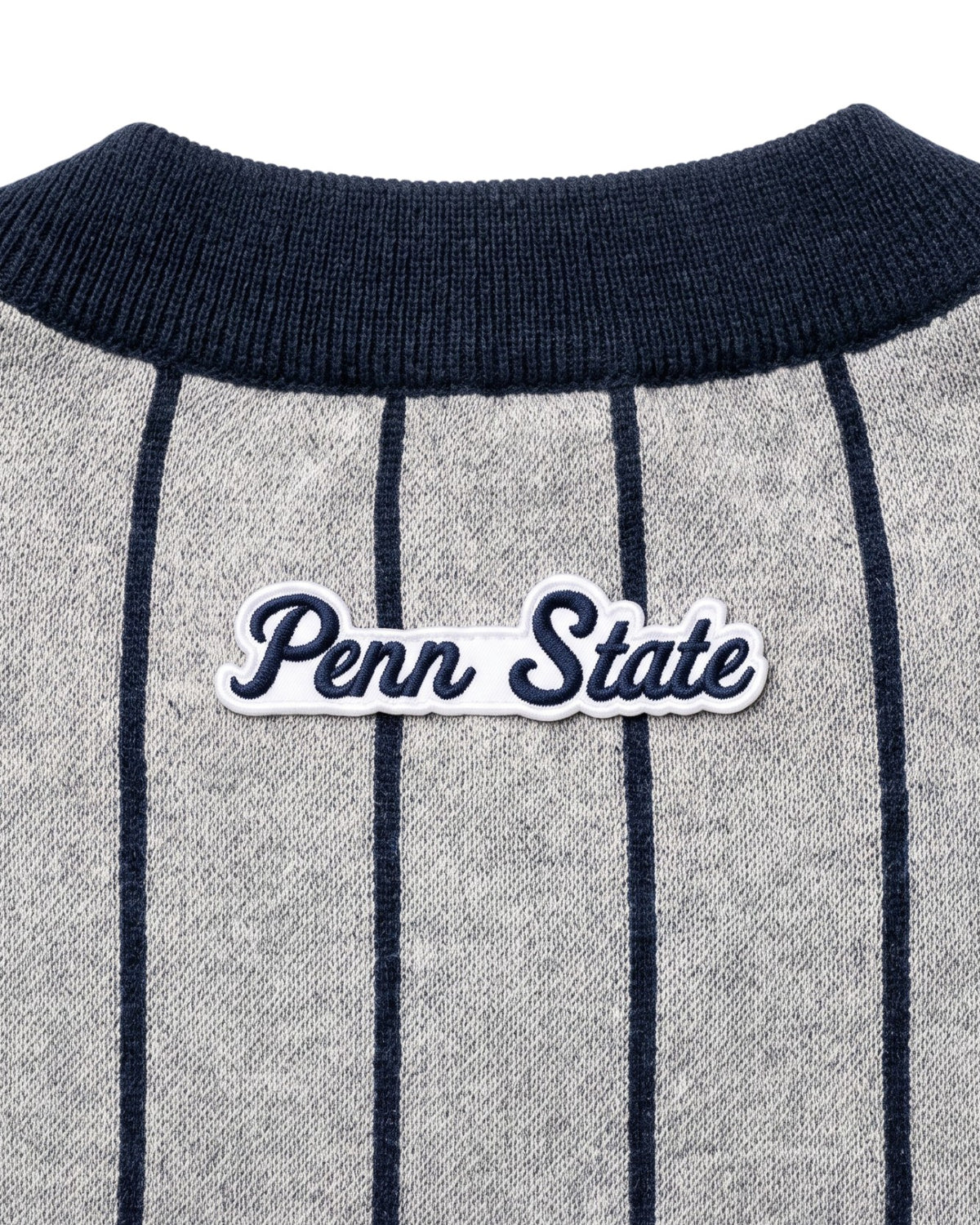 Penn State University script letter crest patch sewn onto upper back below collar of light grey cardigan knit sweater with navy pinstripes and trim.