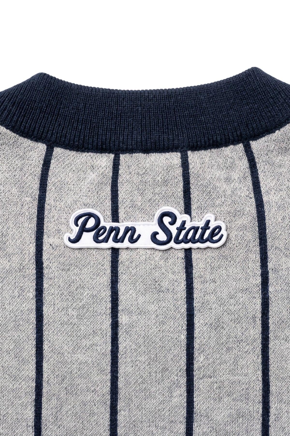 Penn State University script letter crest patch sewn onto upper back below collar of light grey cardigan knit sweater with navy pinstripes and trim.