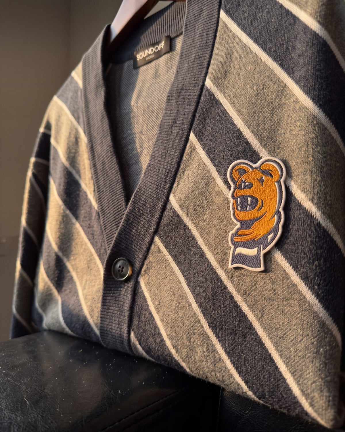Penn State Nittany Lion head embroidered crest patch with scarf in brown, navy and white sewn onto left chest of navy knit cardigan sweater with light grey and white repp stripes.