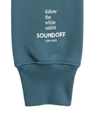 Close-up of the left sleeve on the Heather Teal Bad Bunny Headphones Raglan Sweatshirt, showing the stacked, left-justified "Follow the White Rabbit" text print above the cuff.