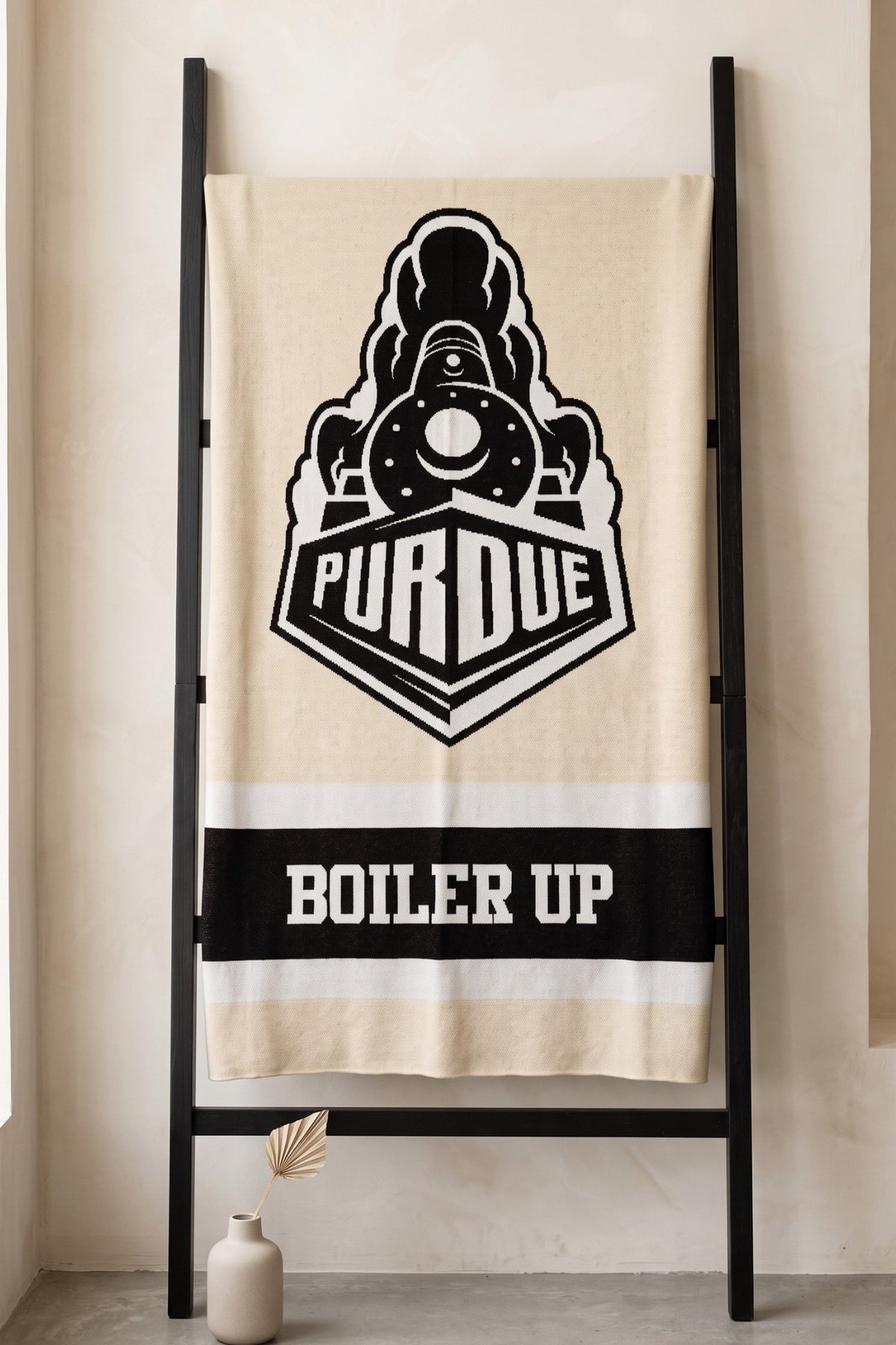 Purdue University varsity knit throw blanket in beige, black, and white with Boilermaker Special graphic by SOUNDOFF hanging on blanket ladder