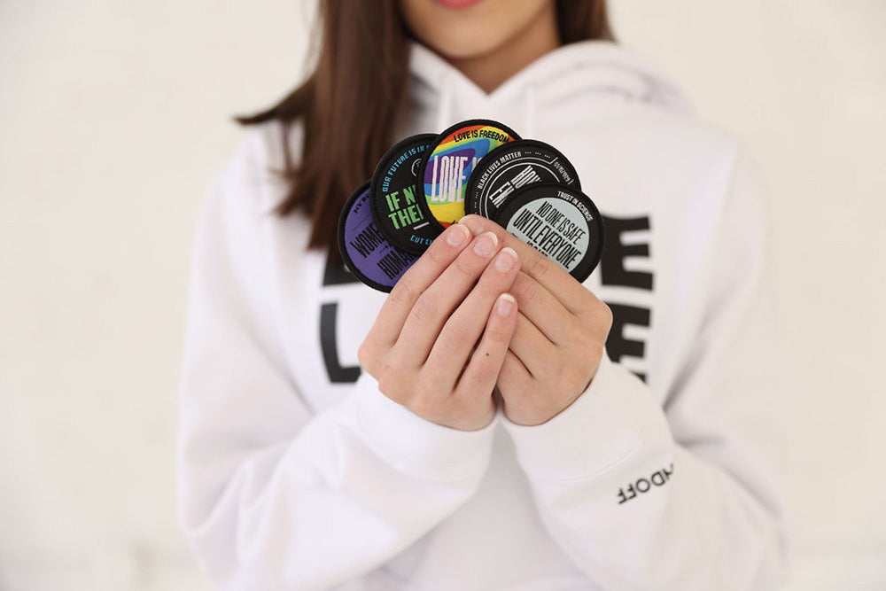 Person wearing Soundoff Love Life Hooded Pullover in White holding five circular Soundoff interchangeable Patches including Love is Love